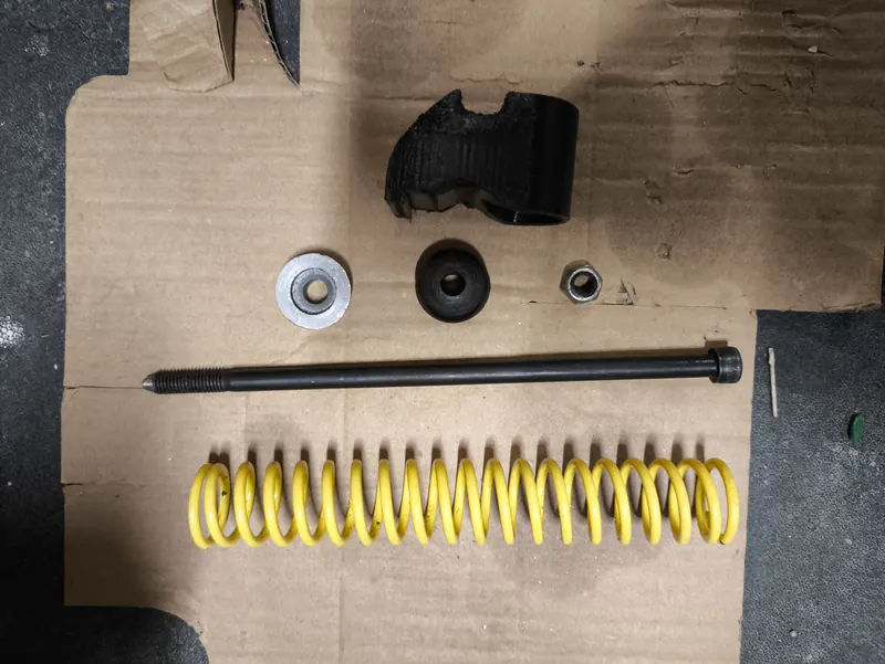 Les pièces du montage posées sur un carton : pièce noire imprimée en PETG, rondelles, longue tige filetée et ressort jaune